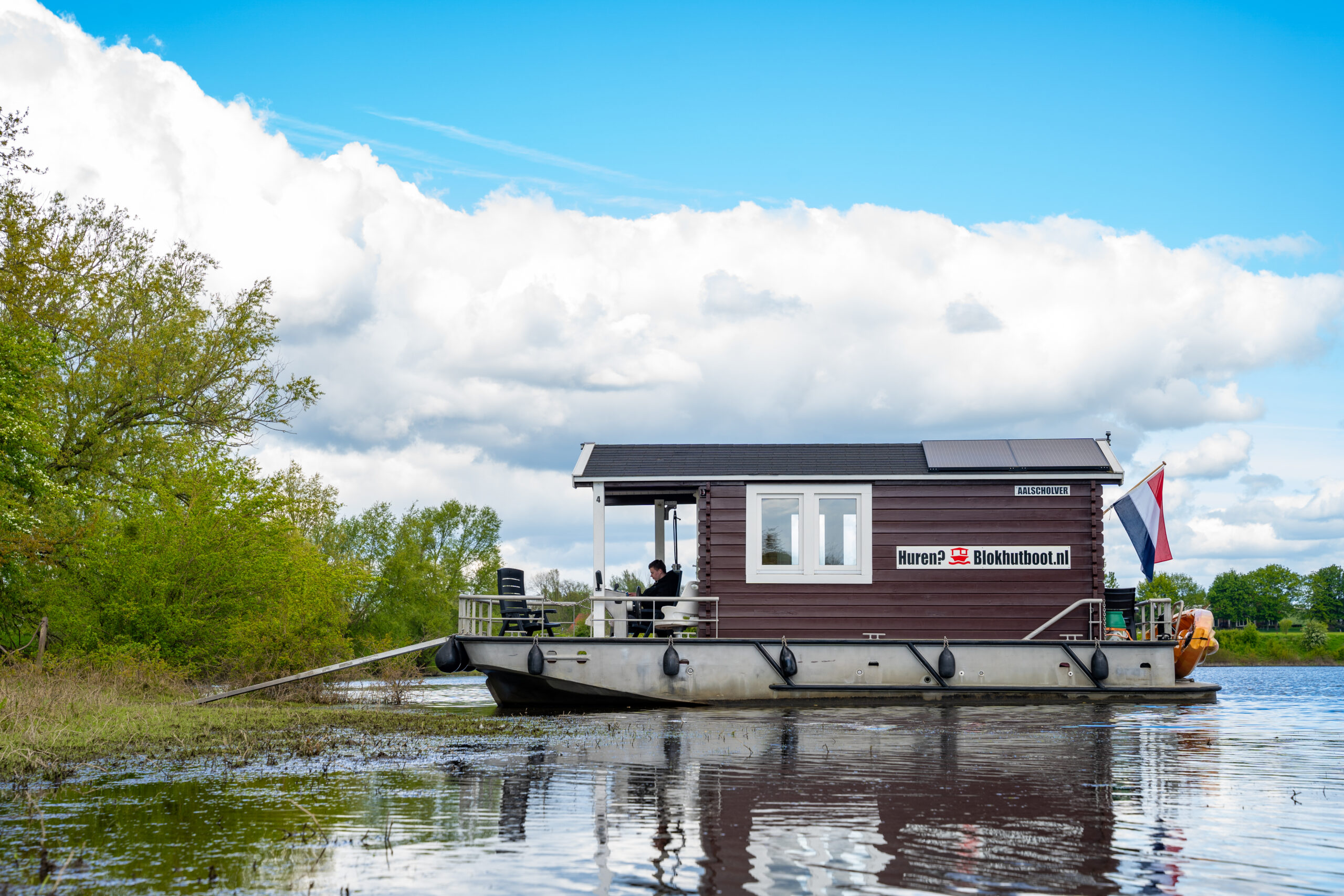 "Een blokhutboot ligt aangemeerd aan de oever van een waterlichaam, omgeven door groene bomen en begroeiing. De boot heeft een houten structuur met een donkerbruine kleur, witte ramen, en een veranda aan de voorkant met stoelen. Op de zijkant van de boot staat de tekst 'Huren? Blokhutboot.nl'. Een Nederlandse vlag hangt aan de achterkant van de boot, en er zijn zonnepanelen op het dak zichtbaar. De lucht is blauw met grote witte wolken."