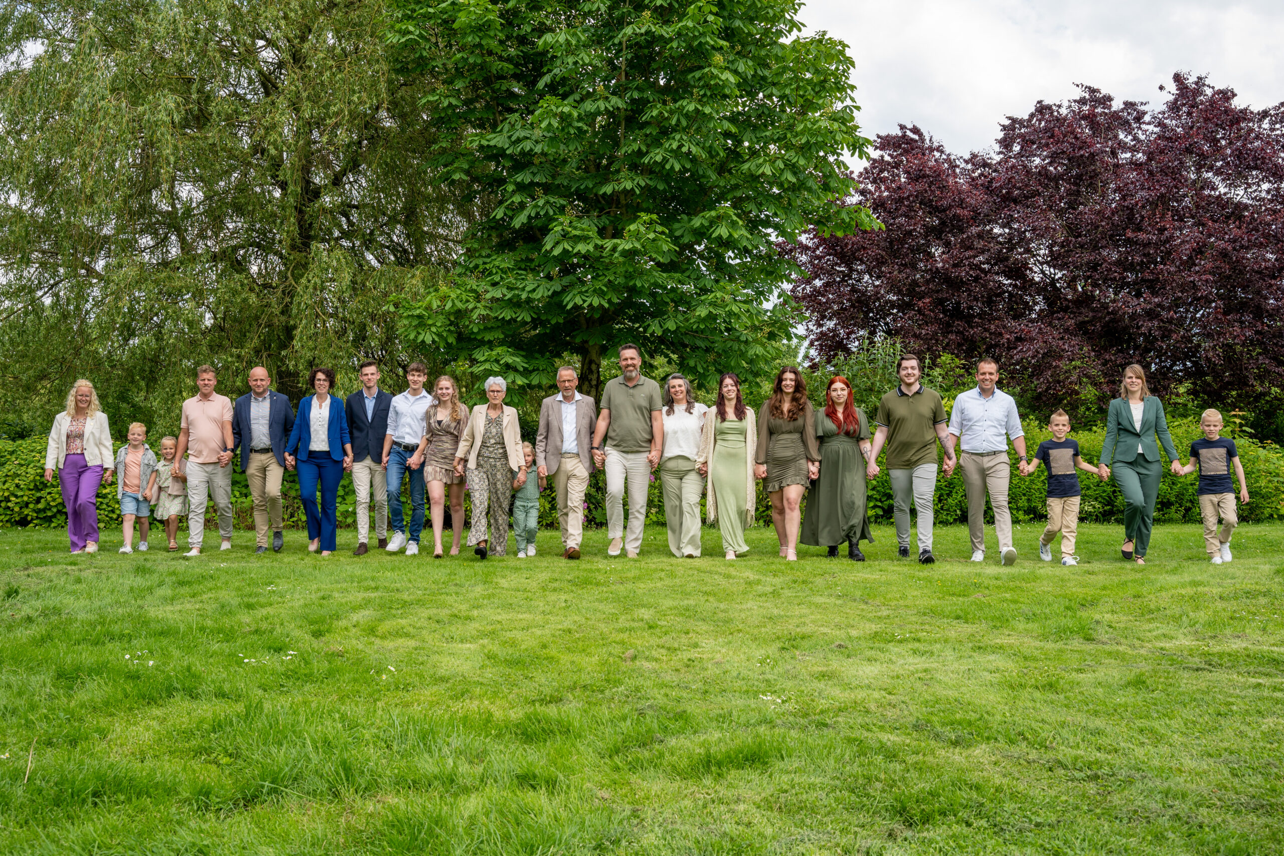 Familiefoto Een grote familie staat hand in hand op een grasveld voor een achtergrond van weelderige bomen. De groep bestaat uit verschillende generaties, waaronder kinderen, volwassenen en ouderen. Iedereen is casual maar netjes gekleed, met groene en aardetinten die veel voorkomen in de kleding. De familie kijkt lachend naar de camera en vormt een rij terwijl ze samen vooruit lopen. Op de achtergrond zijn groene struiken en bomen te zien, wat zorgt voor een natuurlijke en ontspannen sfeer.