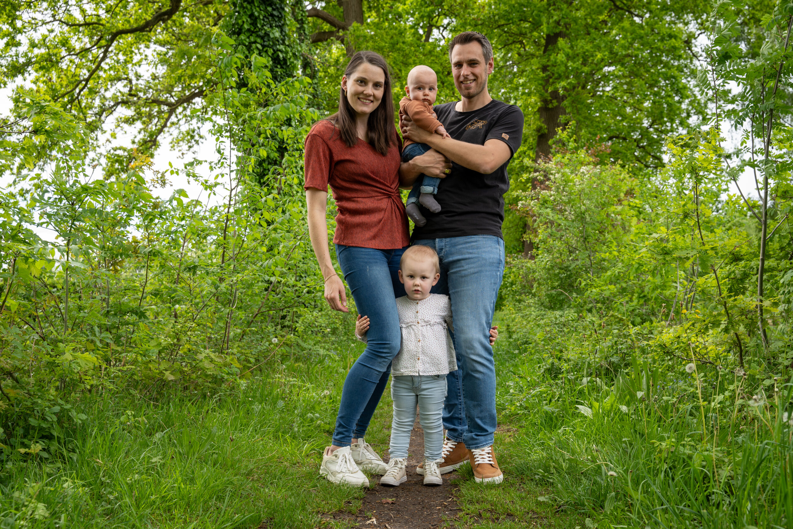 Gezinsfoto Een jong gezin poseert voor een foto in een groen, bosrijk gebied. De moeder draagt een rood T-shirt en spijkerbroek, terwijl ze naast haar peuter staat, die voor haar staat en een witte blouse met lichtblauwe broek draagt. De vader, in een zwart T-shirt en spijkerbroek, houdt een baby in zijn armen. Ze staan op een smal pad omringd door weelderige planten en bomen, wat de foto een natuurlijke, serene sfeer geeft. Iedereen lacht naar de camera, en de familie lijkt ontspannen en gelukkig.