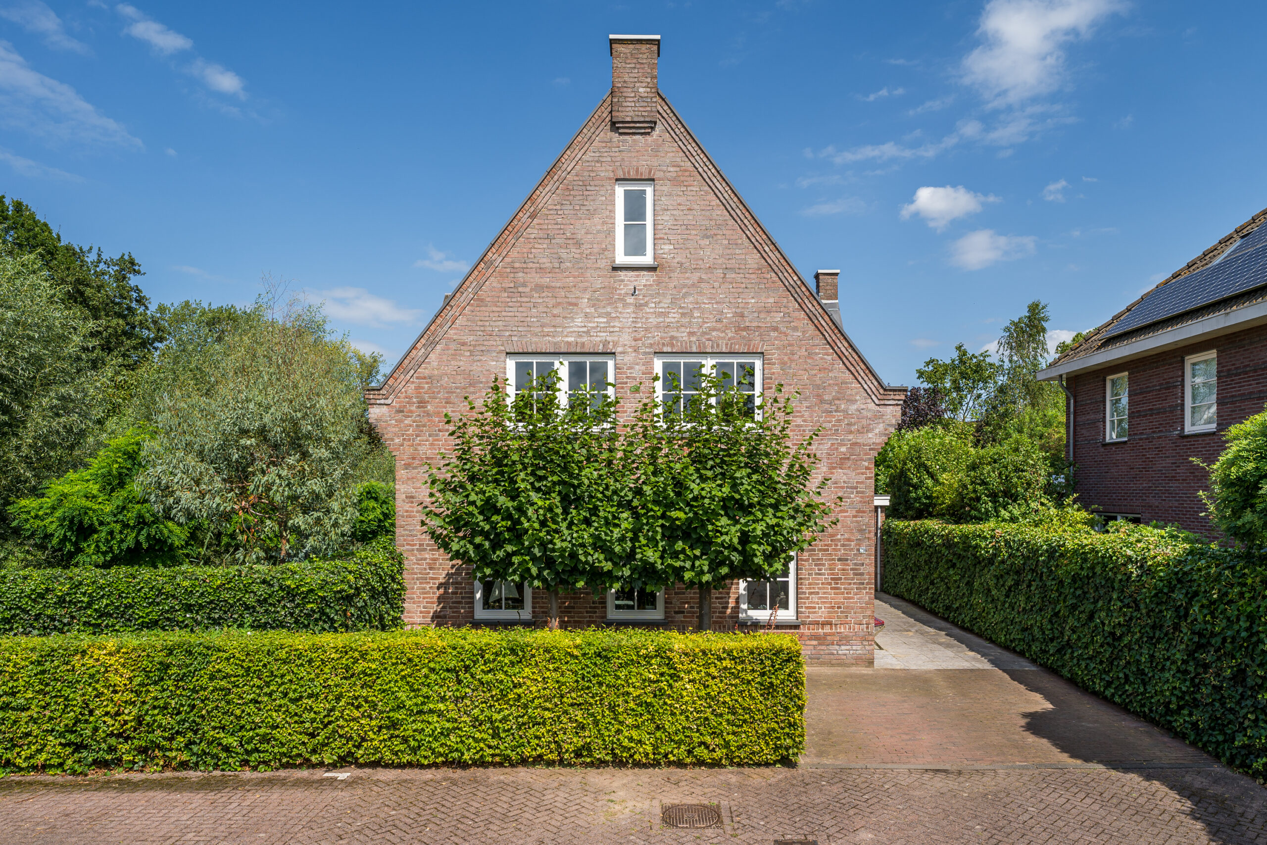 Een bakstenen huis met een puntdak en een schoorsteen, gezien vanaf de voorkant. De gevel heeft twee ramen met witte kozijnen op de begane grond en één raam op de eerste verdieping. Voor de woning staat een rij goed onderhouden struiken en twee bomen met volle groene bladeren die het zicht op de ramen deels blokkeren. Het huis heeft een oprit aan de rechterkant die leidt naar een achterliggende ruimte, en wordt omringd door groene heggen en bomen. De lucht is helderblauw met enkele wolken, wat een zonnige dag suggereert.