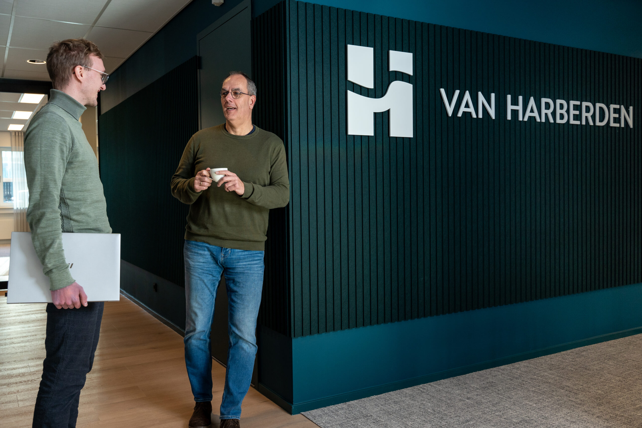 Bedrijfsreportage "Een blokhutboot ligt aangemeerd aan de oever van een waterlichaam, omgeven door groene bomen en begroeiing. De boot heeft een houten structuur met een donkerbruine kleur, witte ramen, en een veranda aan de voorkant met stoelen. Op de zijkant van de boot staat de tekst 'Huren? Blokhutboot.nl'. Een Nederlandse vlag hangt aan de achterkant van de boot, en er zijn zonnepanelen op het dak zichtbaar. De lucht is blauw met grote witte wolken."